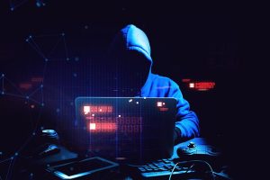 Hooded person with their face obscured sitting in a dark room working on a computer attempting to hack into someones data.